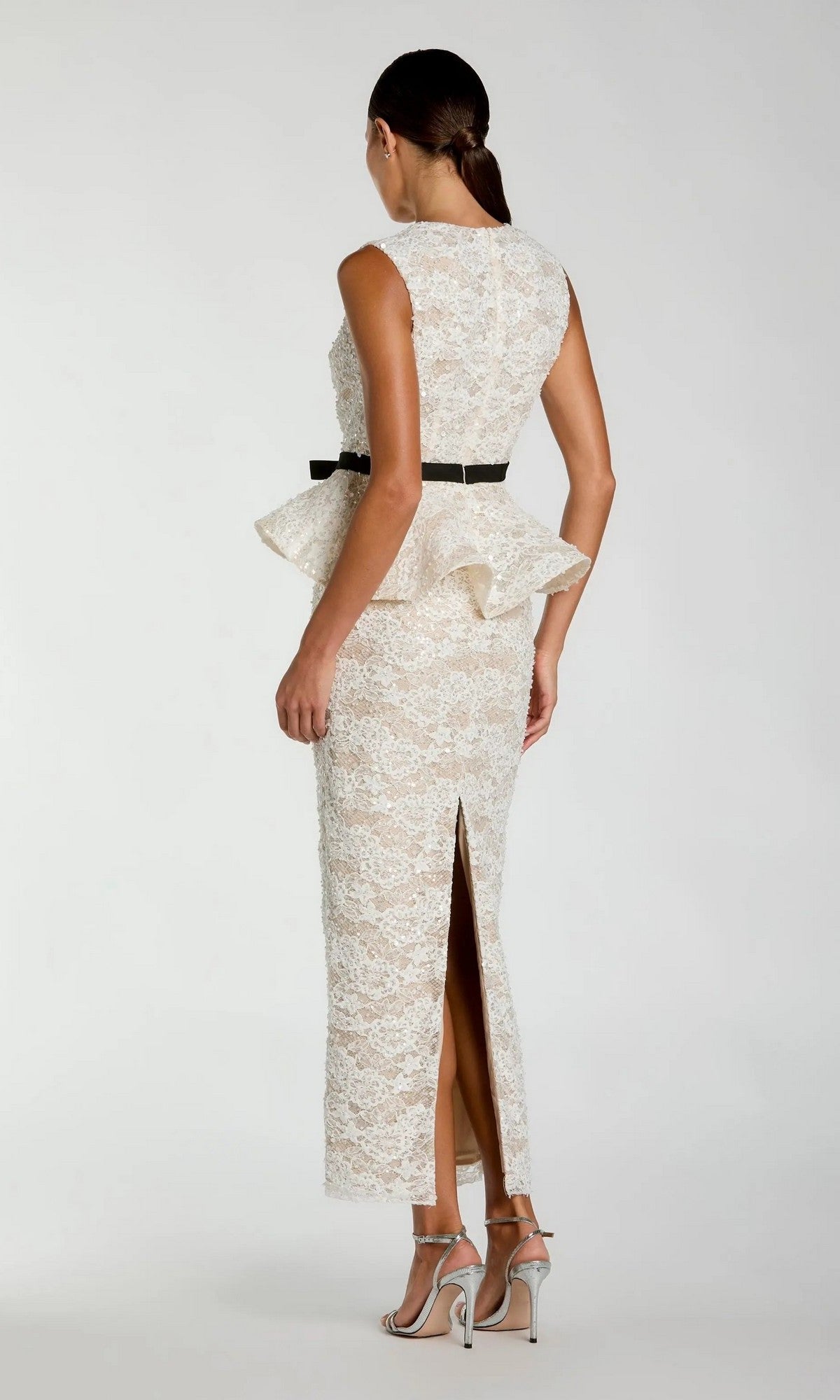 A woman stands in profile against a plain background, wearing the Mac Duggal 21228 long formal dress—an elegant ivory lace gown featuring a peplum waist, black belt, back slit, and silver high-heeled sandals.